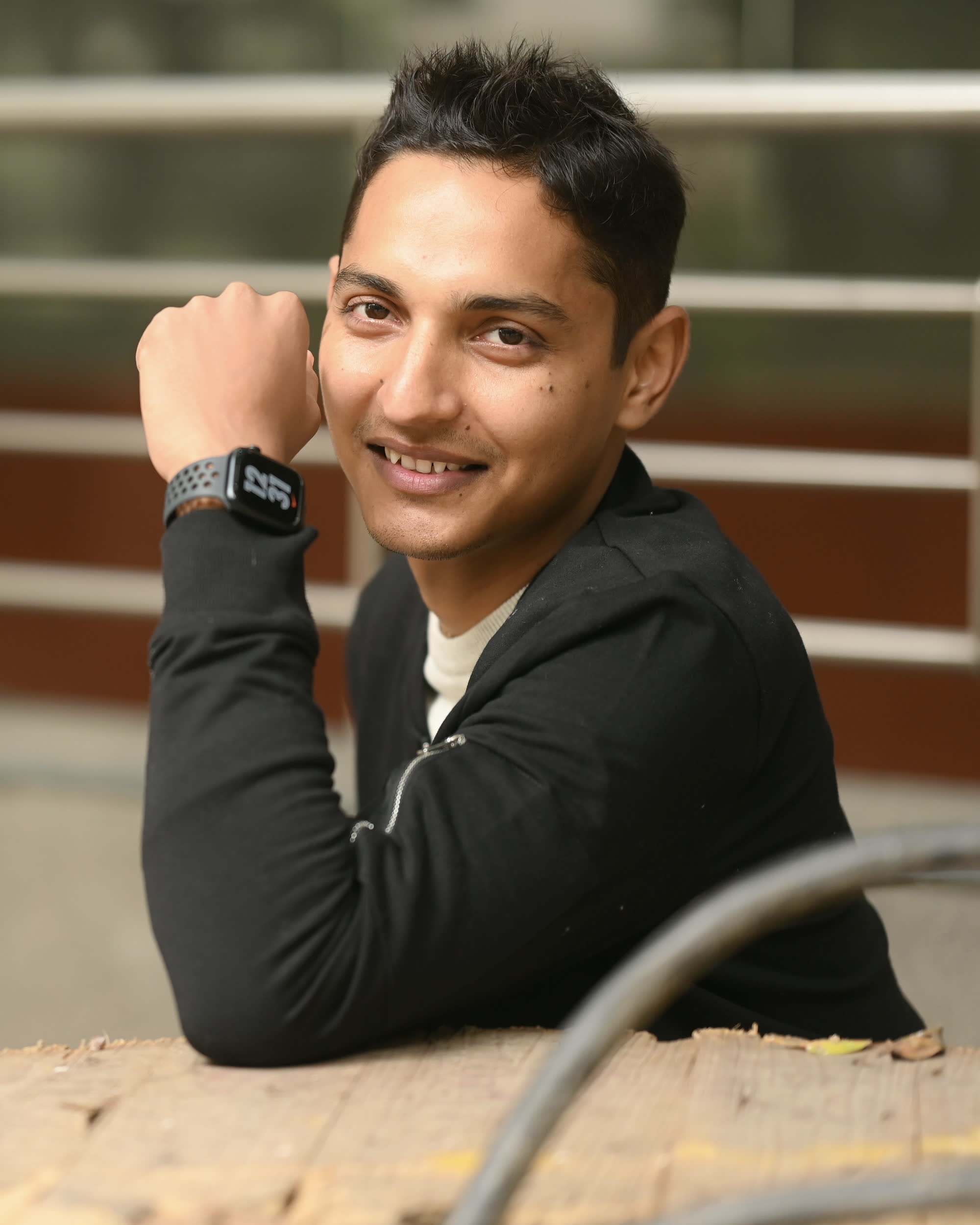 Lokesh outdoors, resting his chin on his hand and smiling slightly off-camera, in soft overcast daylight.